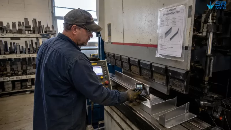 CNC press brake work area showing a freshly bent stainless steel L-bracket on the machine table with back gauge fingers, V-die groove, digital angle readout displaying 90.1 degrees, and a tooling rack with color-coded punch and die sets in the background