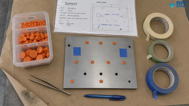 Flat-lay photograph of a steel enclosure panel with orange silicone masking plugs in threaded holes, surrounded by masking tape rolls, tweezers, and a work order with masking instructions during powder coating preparation