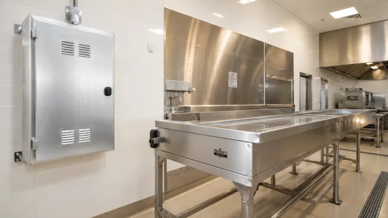 A modern commercial kitchen featuring a stainless steel food processing table and a wall-mounted aluminum electrical enclosure, demonstrating real-world material applications